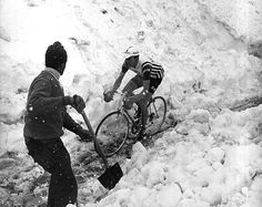 black and white photograph of two men on bicycles in the snow, one pushing another bike up a snowy hill