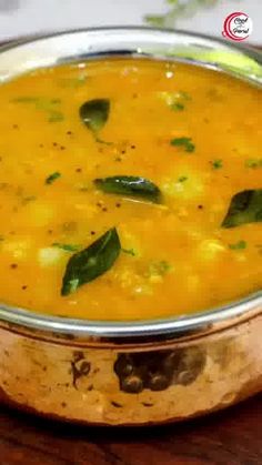 a metal bowl filled with yellow soup on top of a wooden table next to a spoon