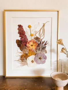 an arrangement of dried flowers in a white frame on a wooden table next to a vase