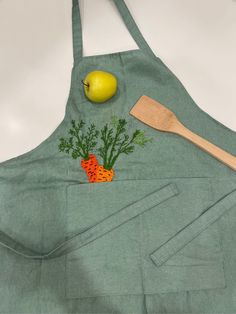 an apron with carrots, apples and parsley embroidered on the front is next to a wooden spoon