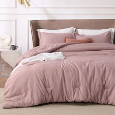 a bed with pink comforter and pillows in a white room next to a lamp