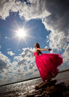 a woman in a pink dress is standing on rocks near the water and looking up into the sky