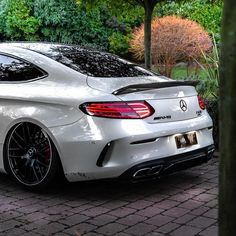 the rear end of a white car parked in front of some trees and bushes on a brick road