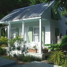 a small white house with green shutters on the front and side windows, surrounded by greenery