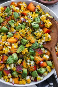 a bowl filled with corn, avocado, and other veggies next to a wooden spoon