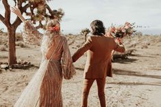 a man and woman walking in the desert with flowers on their head, holding hands