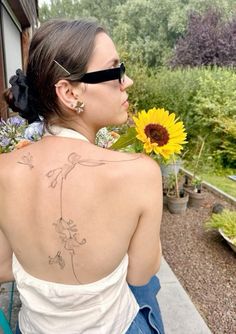 a woman with a sunflower tattoo on her upper back and right arm is sitting in front of a house