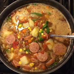 a person holding a spoon over a bowl of soup with sausage, potatoes and corn