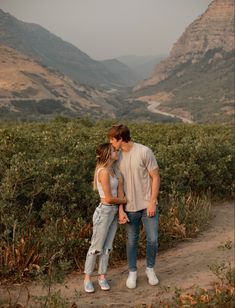 a man and woman are kissing on a path in front of some hills with trees