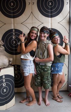 three young people posing for a photo in front of an archery target wall with arrows on it