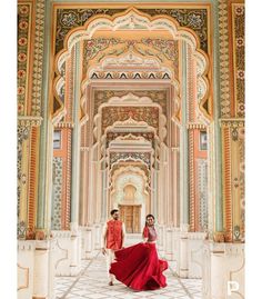 two women in red dresses are walking through an ornate building with columns and arches on either side