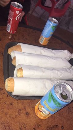 four hot dogs wrapped in white paper on a table with cans of soda and can