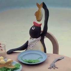 an animated penguin sitting at a table with food on it's plate and another animal standing next to him