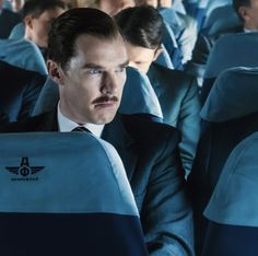 a man in a suit and tie sitting on an airplane with other people behind him