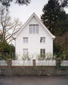 a white house sitting next to a lush green tree filled forest in front of a tall white fence