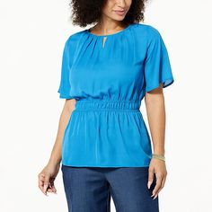 a woman with curly hair wearing a bright blue blouse and jeans, standing in front of a white background