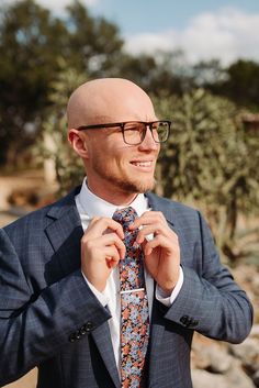 a bald man wearing glasses and a suit is straightening his tie with one hand while standing in front of some rocks