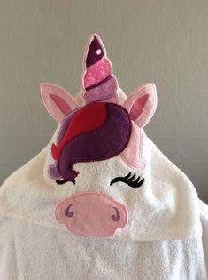 a white towel with a pink and purple unicorn on it's face, sitting next to a gray wall