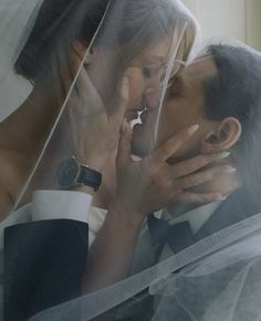 a man and woman kissing under a veil in front of a window with their hands on each other's face