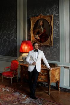 a man in a white tuxedo standing next to a table with a painting on it