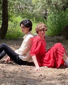 two people sitting on the ground in front of some trees and grass with one person wearing a red dress