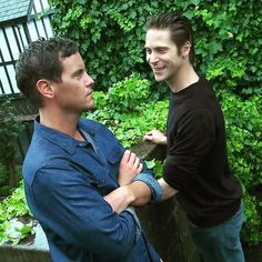 two men standing next to each other in front of green bushes and trees, one with his arm around the other's neck