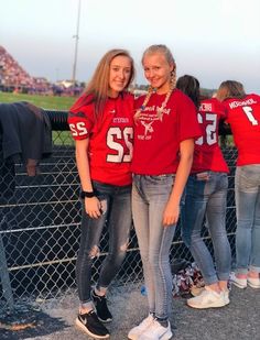 two girls in red shirts standing next to each other near a fence and some fans