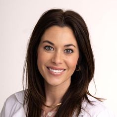 a woman with long brown hair wearing a white shirt and gold necklace smiling at the camera