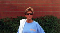 a woman in sunglasses and a blue shirt is holding a skateboard near a brick wall