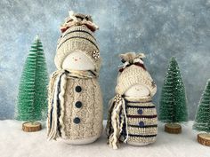 two knitted snowmen standing next to each other in front of small christmas trees