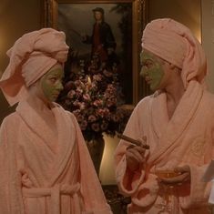 two women in pink robes with facial masks on and one is holding a spoon to her mouth