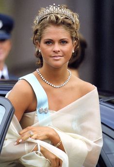 a woman in a white dress and tiara standing next to a car with a man in uniform behind her