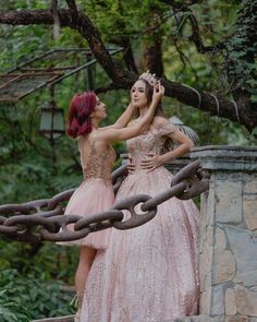 two women dressed in pink dresses standing on a bridge with chains hanging from it's sides