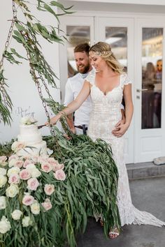 a bride and groom cutting their wedding cake at the reception table with greenery in front of them