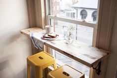 two yellow stools sitting in front of a window next to a small table with a vase on it