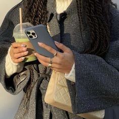 a woman is holding a drink and looking at her cell phone while wearing a gray coat