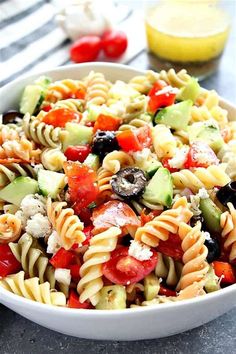 a pasta salad in a white bowl on a table with tomatoes, cucumbers and olives