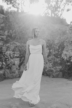 black and white photograph of woman in dress walking on beach near rocks with sun shining through trees