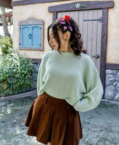 a woman wearing a green sweater and brown pleated skirt with flowers in her hair