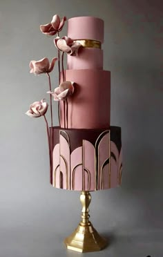 a three tiered pink and gold cake with flowers on the top, sitting on a pedestal