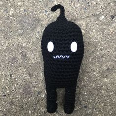 a crocheted black cat with white eyes and nose sitting on the ground next to a sidewalk