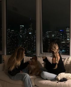 two women sitting on a couch looking out the window at the city lights in the distance