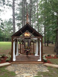 a wooden gazebo with white drapes and flowers in the center surrounded by trees