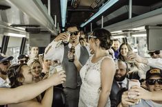 a bride and groom drinking beer on a bus with their wedding party in the background