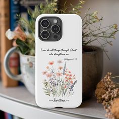 a white phone case with flowers and bible verse on the back, sitting on a shelf next to a potted plant