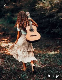 a woman in a dress is holding a guitar and walking through the woods with her back to the camera