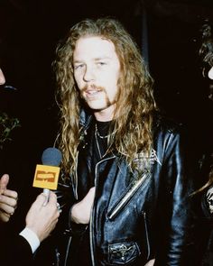 a man with long hair wearing a black leather jacket talking to someone in front of him