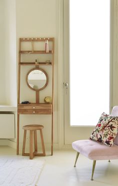 a pink chair sitting in front of a window next to a mirror and table with a vase on it