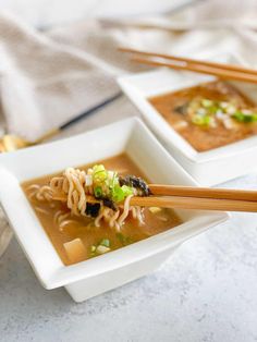 two white square bowls filled with soup and chopsticks on top of a table