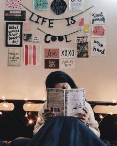 a woman sitting on a bed reading a book in front of a wall covered with posters